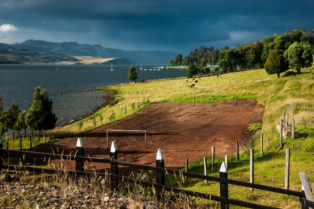 Hillside paddock with wooden fence overlooks a scenic bay and mountains under dramatic sky.