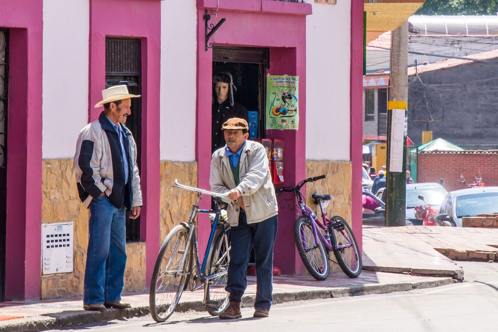 People walk past pink painted buildings and storefronts along a narrow cobblestone street in a Latin American town.