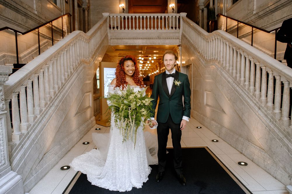 Elegant wedding procession in grand marble staircase venue with ornate chandelier and white balustrades.