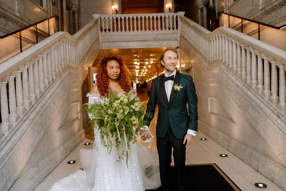 Wedding party poses on elegant grand white staircase in formal attire with emerald green bridesmaid dresses.