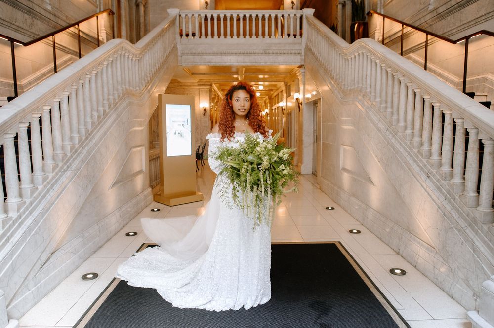 Wedding party members walking down an ornate white staircase in a grand venue with crystal chandeliers.