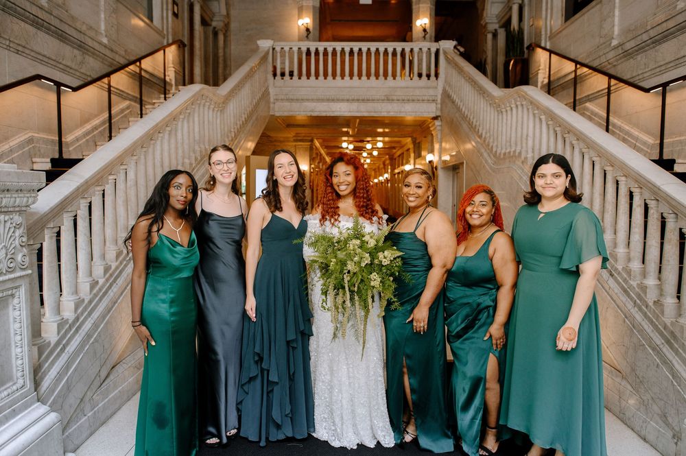 A group wearing dark teal and navy blue dresses stands together on a grand marble staircase in an elegant setting.