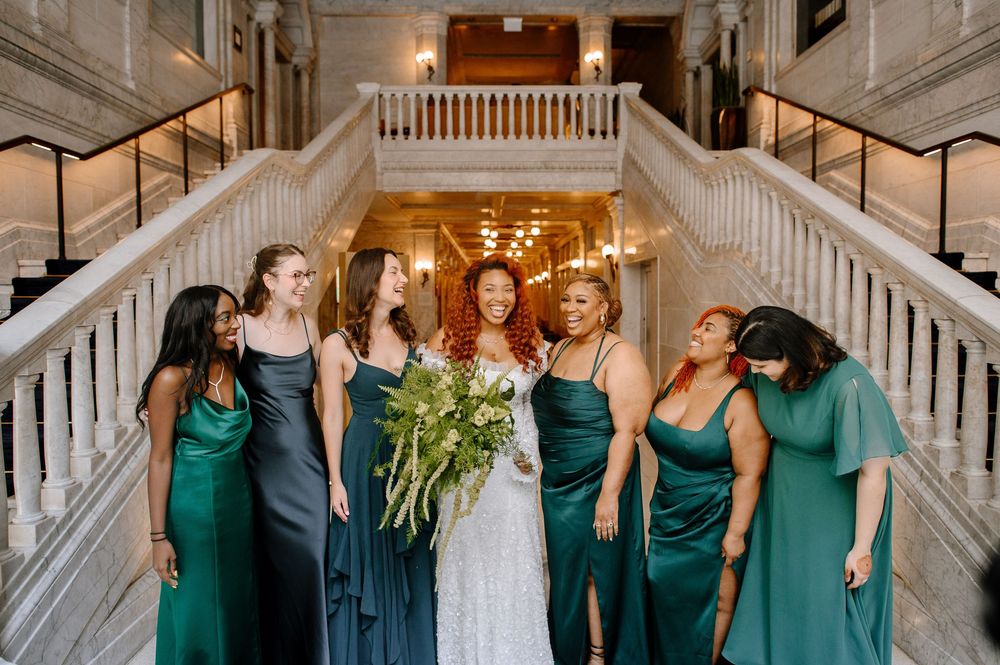 Bridesmaids in emerald green dresses gather around the couple on a classic white marble staircase.