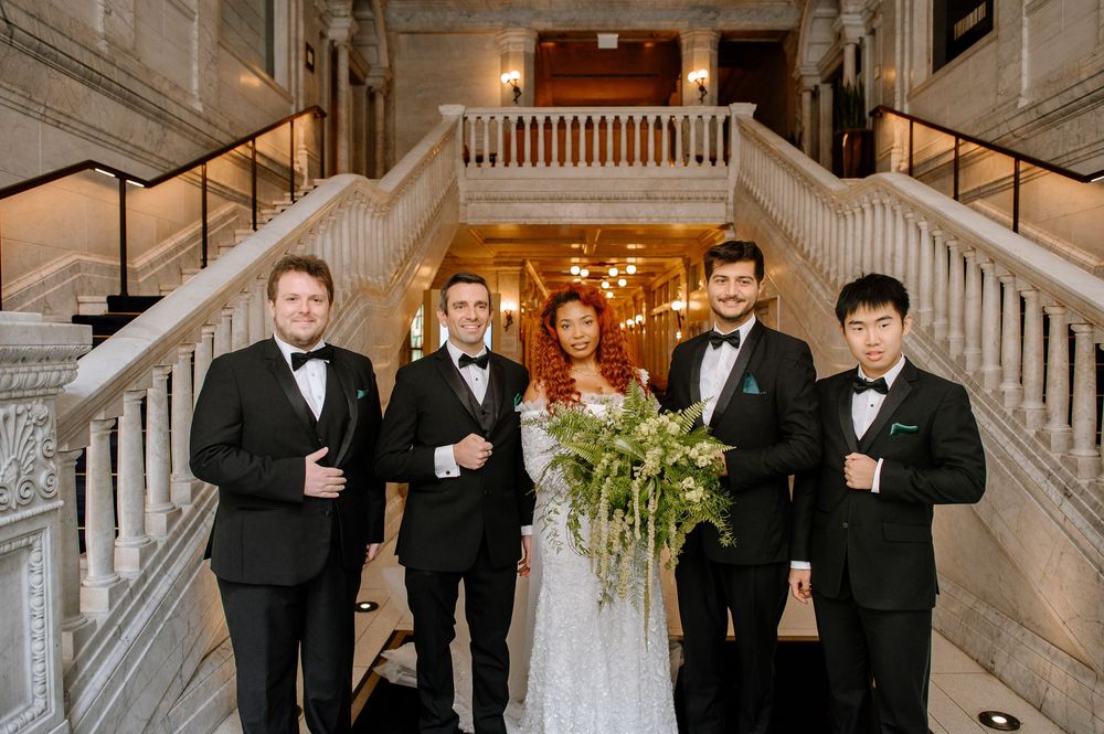 Wedding party members in black tuxedos and formal wear positioned along a luxurious white marble staircase.