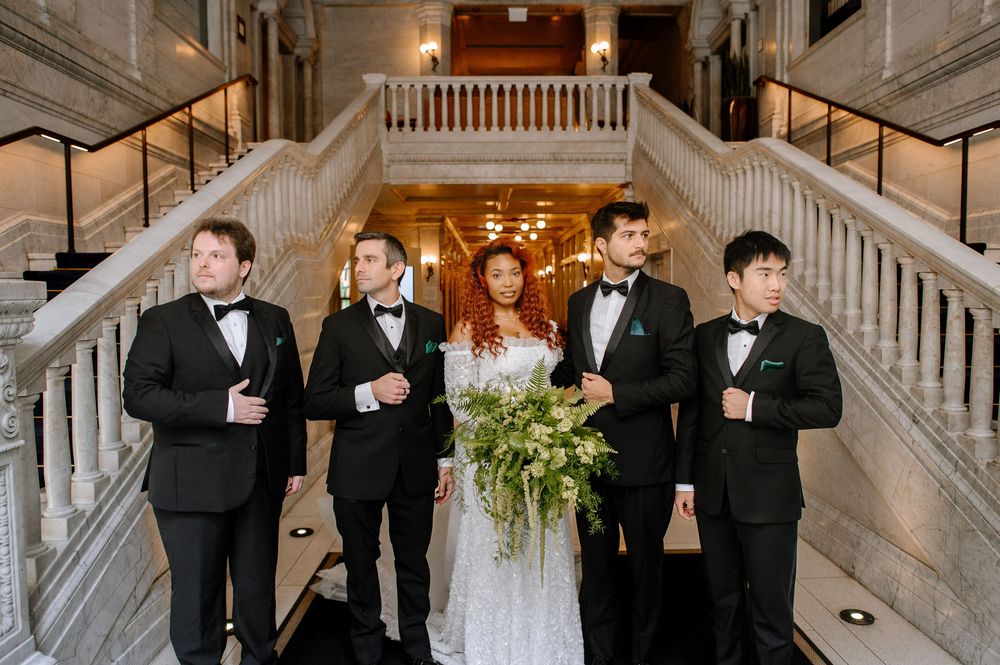 Wedding party poses elegantly on grand marble staircase in formal black tuxedos and bridal attire.