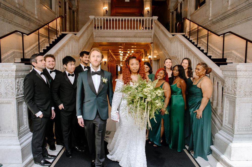 Wedding party poses on an elegant marble staircase in a grand venue with chandelier lighting overhead.