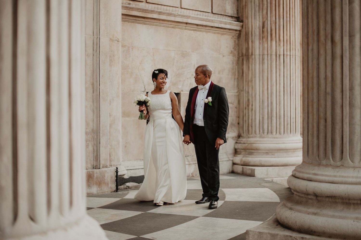 Bride and groom hold hands walking between columns at St Pauls Cathedral. Bride in white strapless gown, groom in black suit