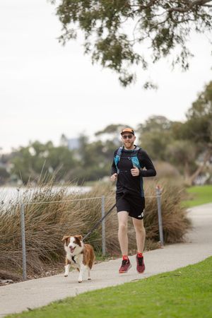Runner in black athletic wear jogging on path with brown and white dog alongside waterfront.