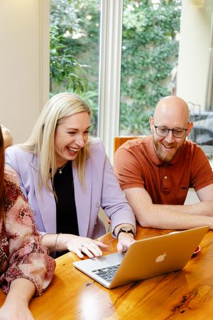 A collaborative work session shows two people sharing a laugh while looking at a laptop on a wooden table.