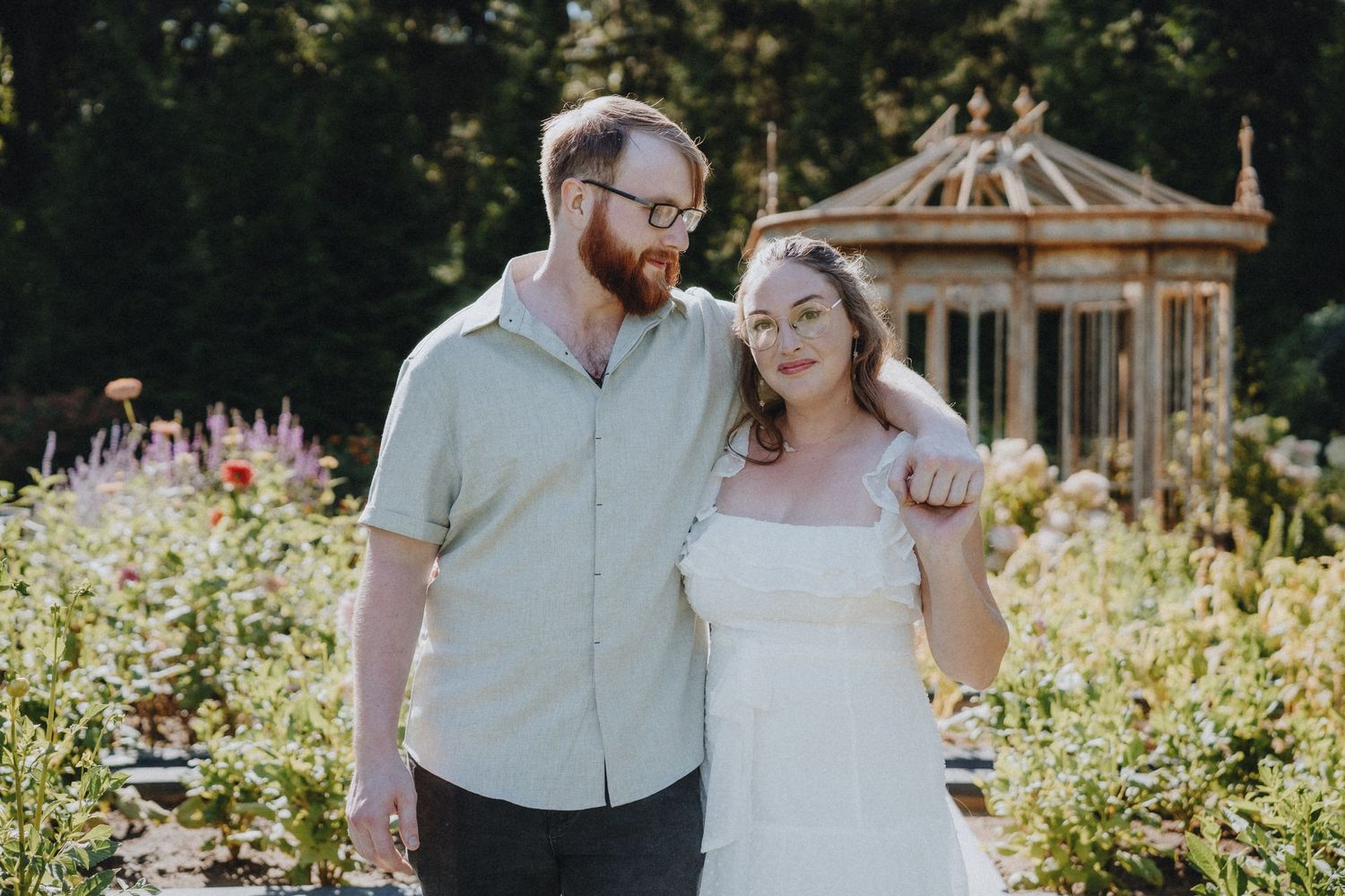 Happy couple posing in front of an ornate garden gazebo surrounded by blooming flowers.