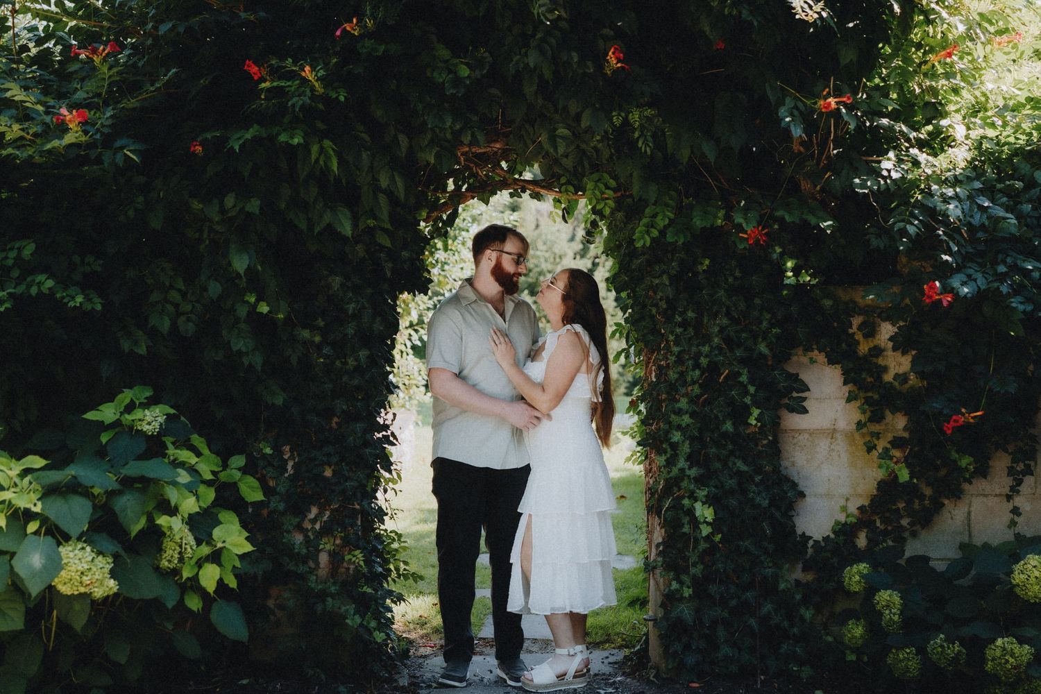 Couple standing beneath a romantic ivy-covered archway with red flowers.