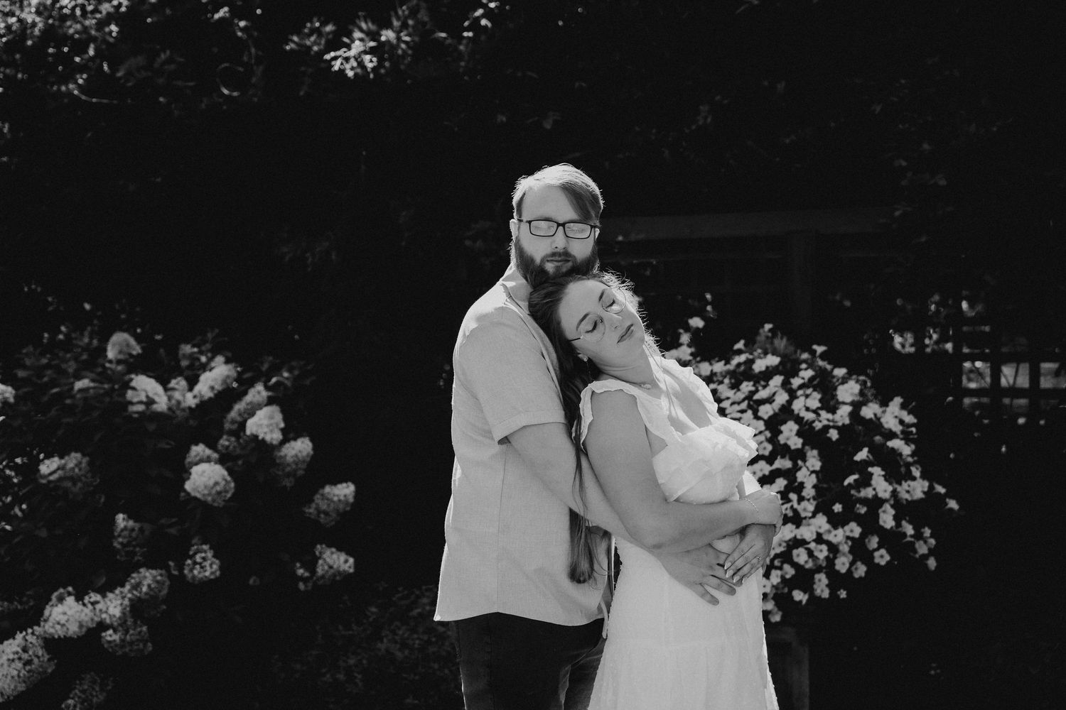 Black and white portrait of a couple embracing in a garden with blooming white flowers.