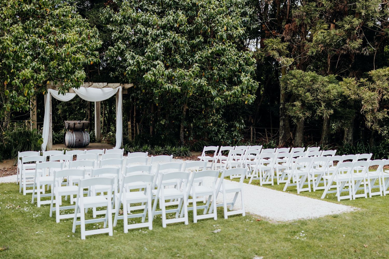 White chairs and a decorative arch are arranged for an outdoor wedding ceremony in a lush garden setting.