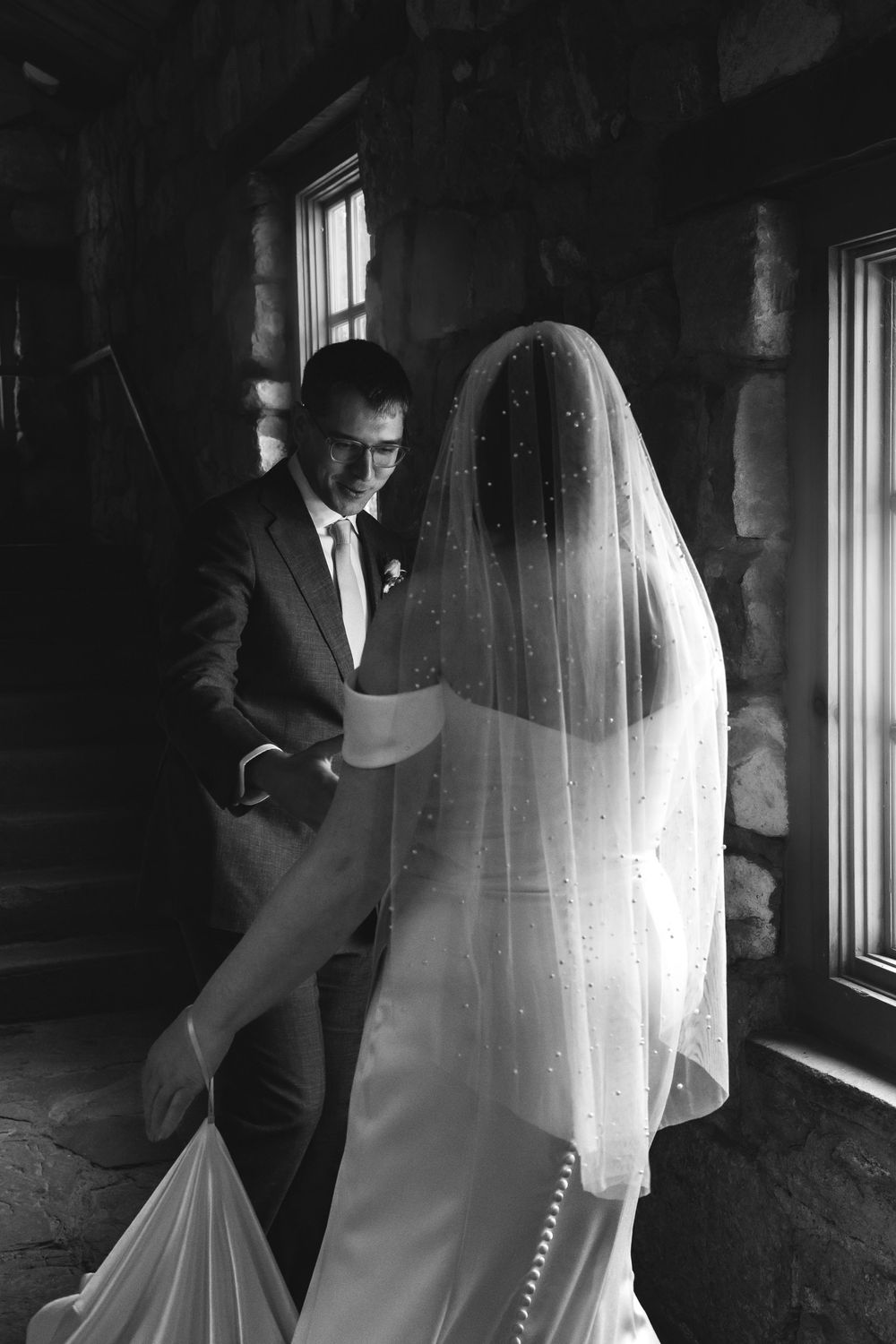 Black and white photo of groom in suit lifting bride's pearl-dotted veil during intimate first look in stone building.