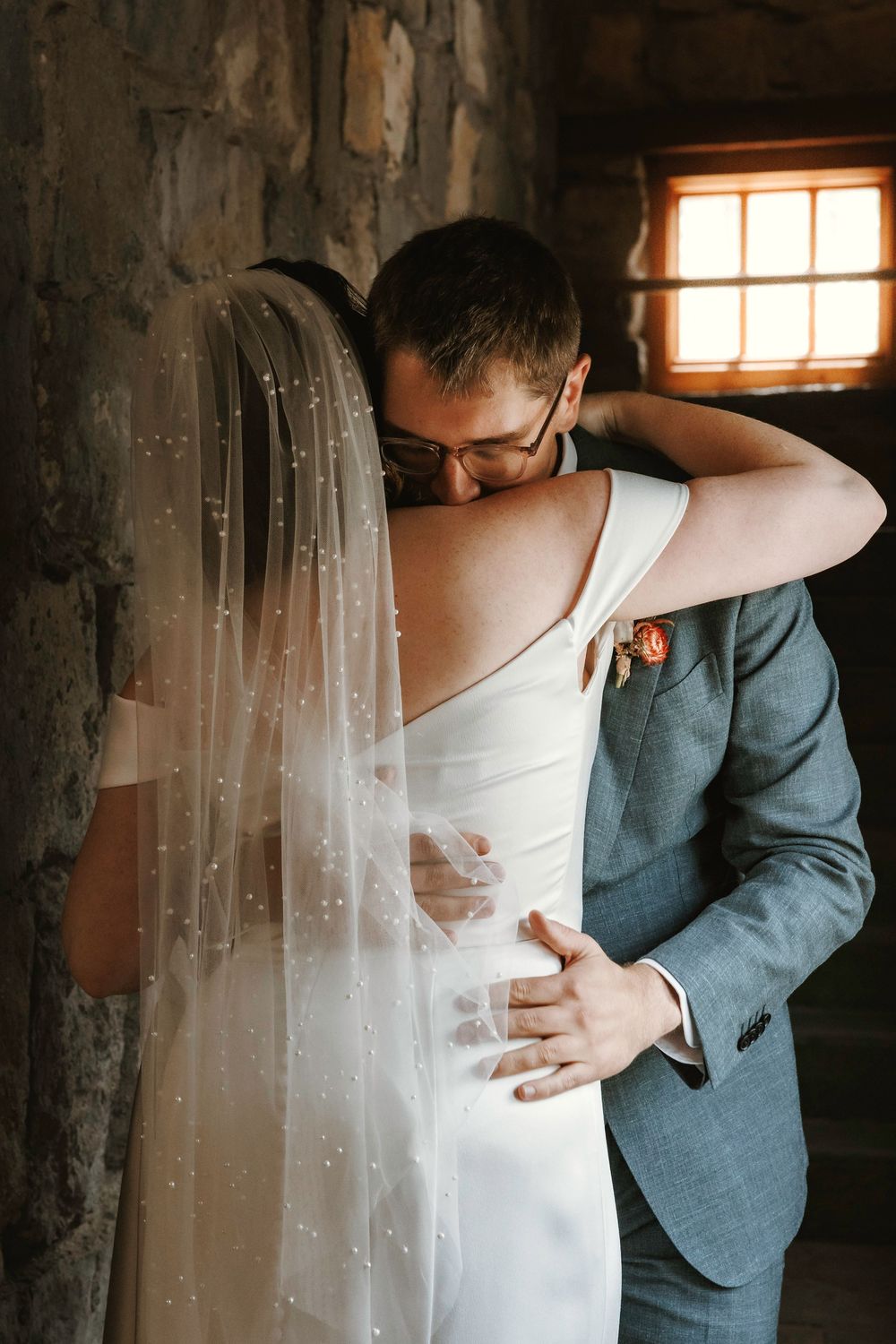 Groom in blue suit embracing bride in off-shoulder white gown with pearl veil during emotional wedding first look moment.
