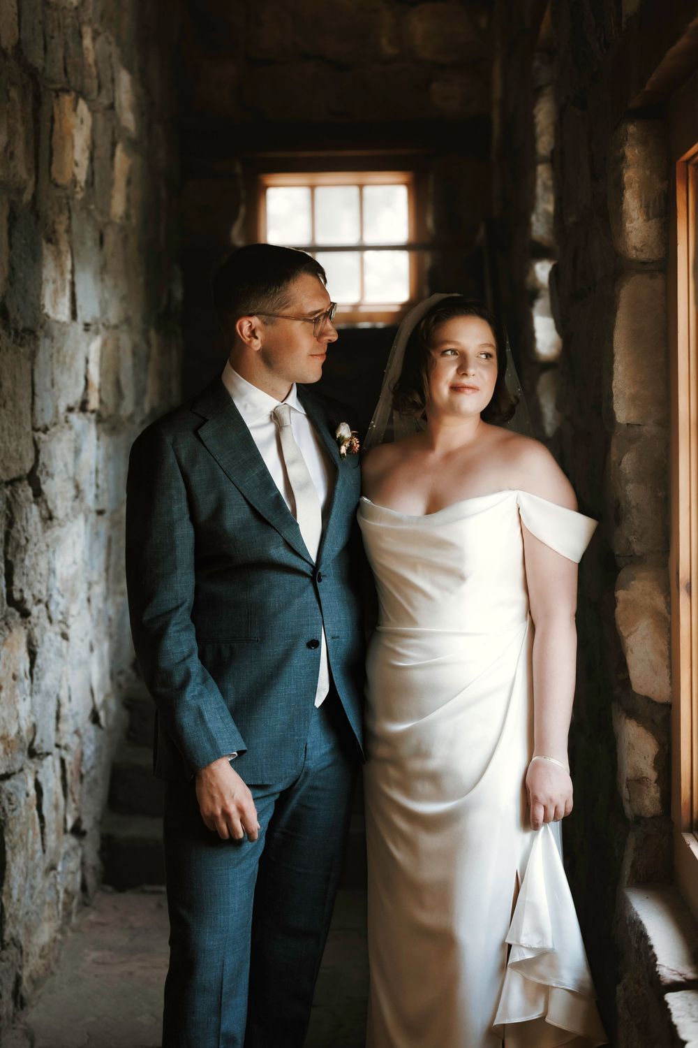 Bride in off-shoulder white satin gown and groom in teal suit posing together in rustic stone building with wooden windows.