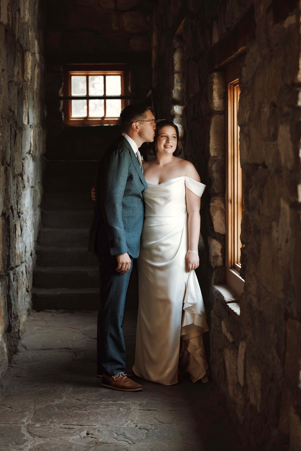 Groom kissing bride on cheek as she smiles in elegant white satin dress inside a romantic stone stairwell venue.