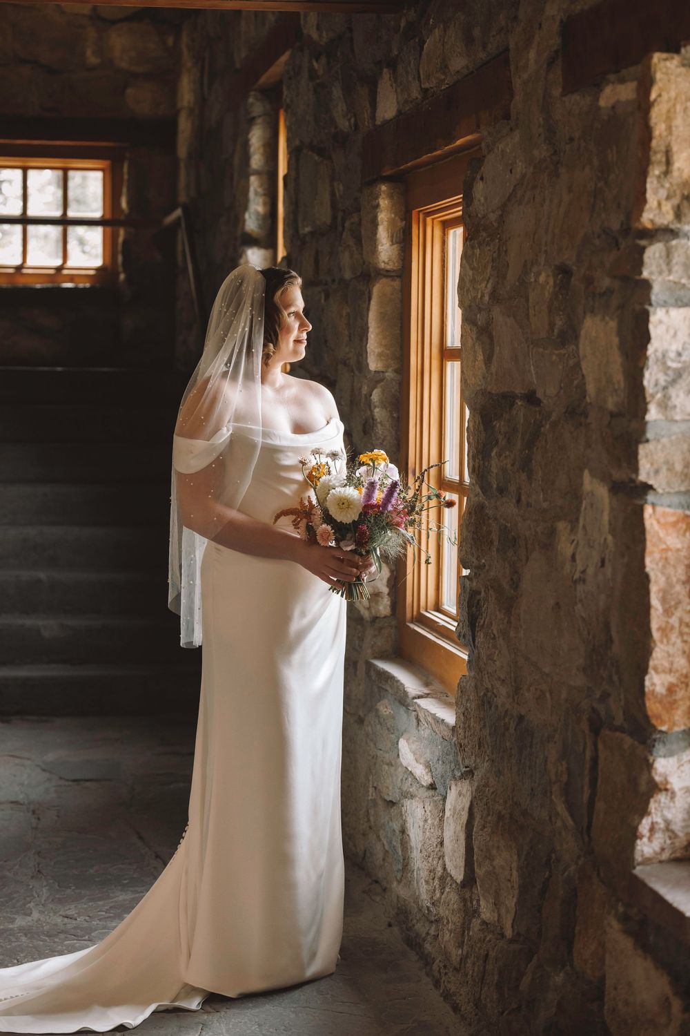 Bride in off-shoulder white gown and veil holding colorful wildflower bouquet while gazing out wooden window in stone venue.