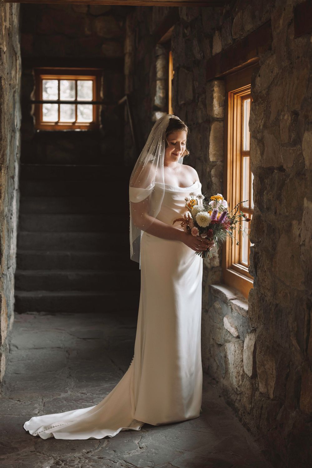 Bride in fitted white satin off-shoulder gown with veil admiring her colorful bridal bouquet by a warm wooden window.