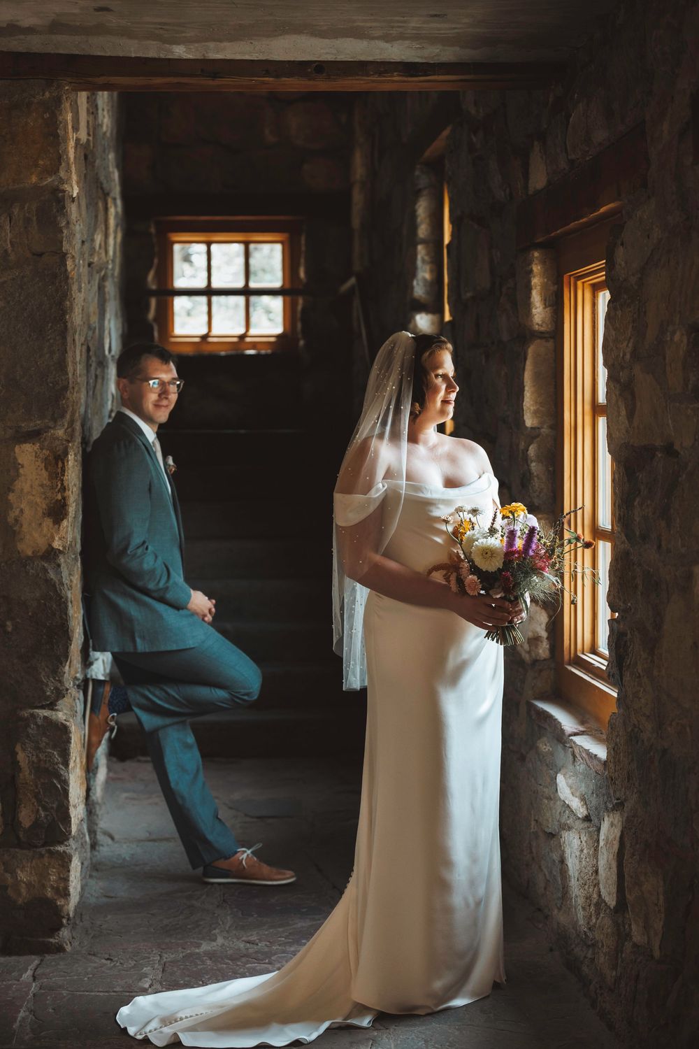 Bride in white gown with veil holds bouquet by window while groom in teal suit sits on stairs in rustic stone venue.