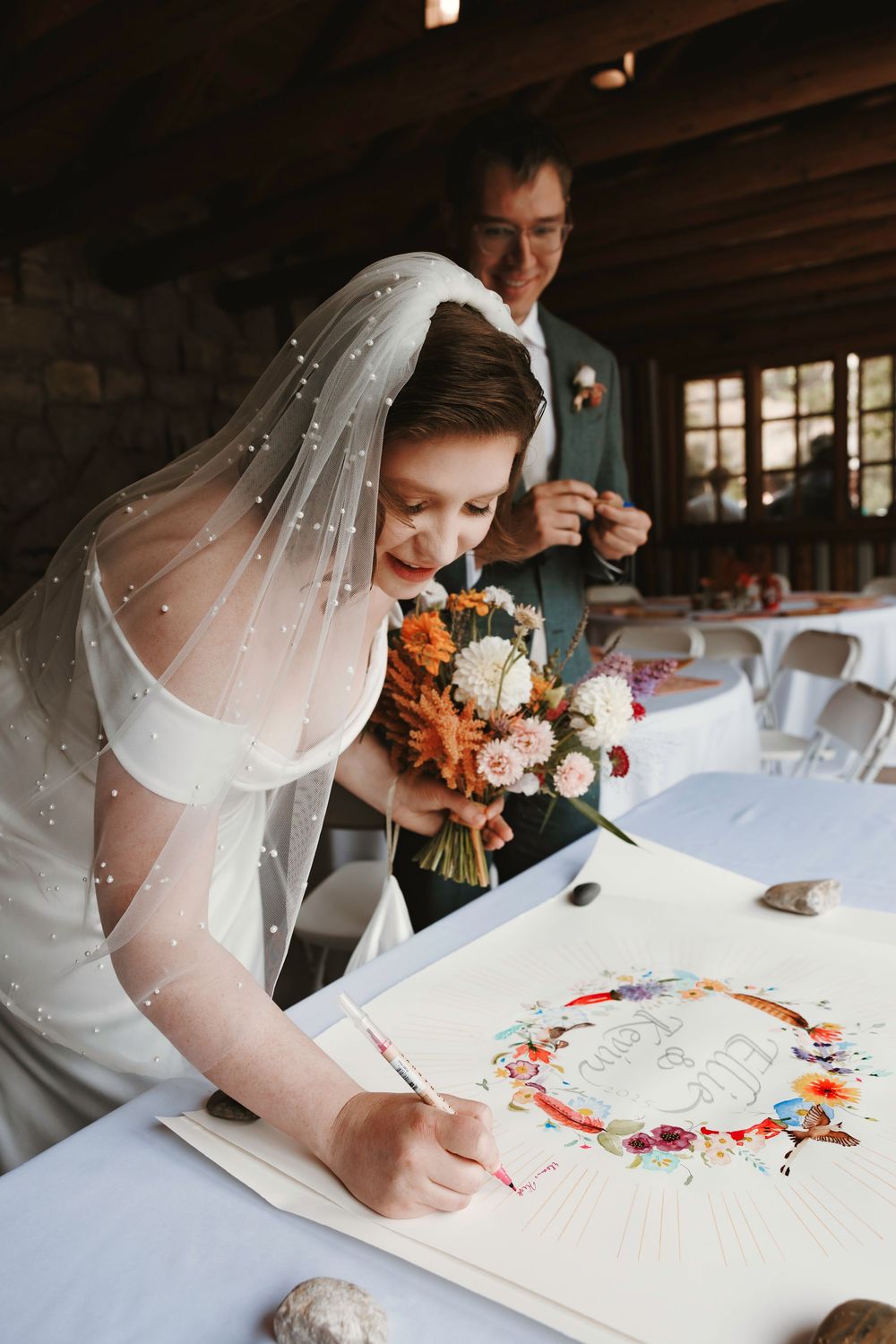Bride in pearl veil signs wedding registry document with colorful floral wreath design while groom stands behind her.