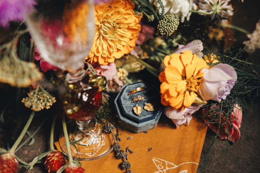 Colorful wedding flat lay with orange zinnias, pink roses, ring box with gold rings, and vintage perfume bottle on golden fabric.