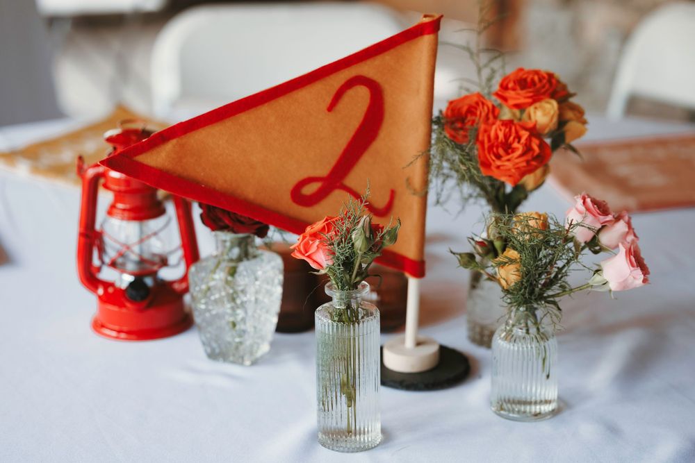 Wedding table centerpiece with orange pennant number two flag, red lantern, and small bud vases with pink and orange flowers.