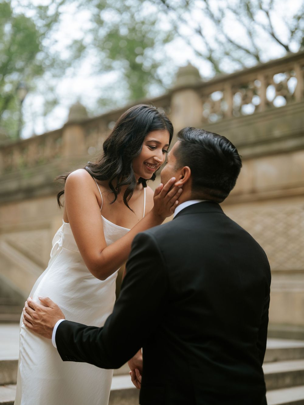 A romantic moment between newlyweds on stone steps with an ornate railing and trees in the background.