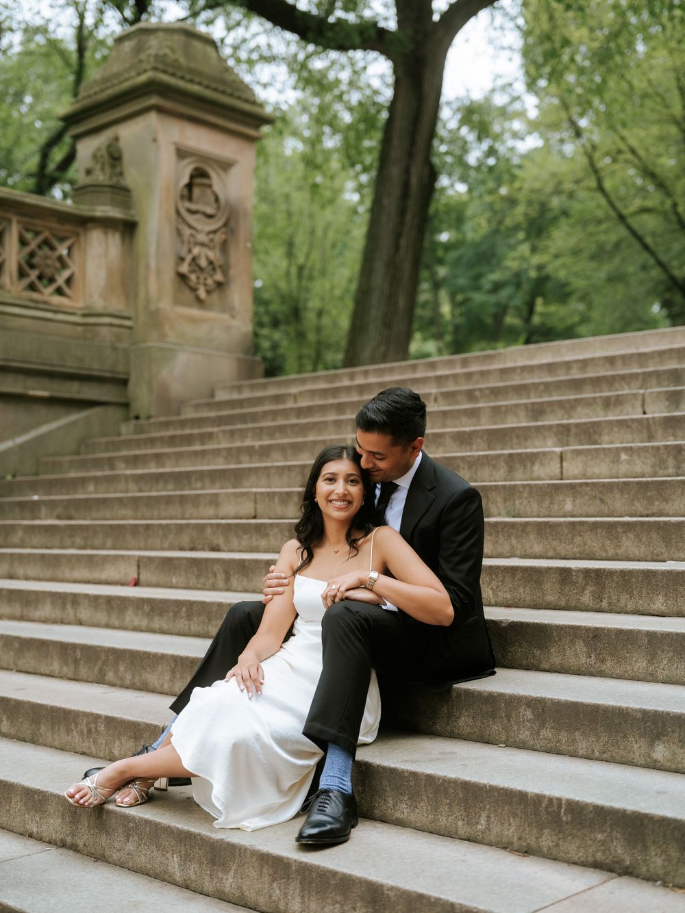A romantic couple shares intimate moments on stone steps surrounded by lush greenery in a park setting.