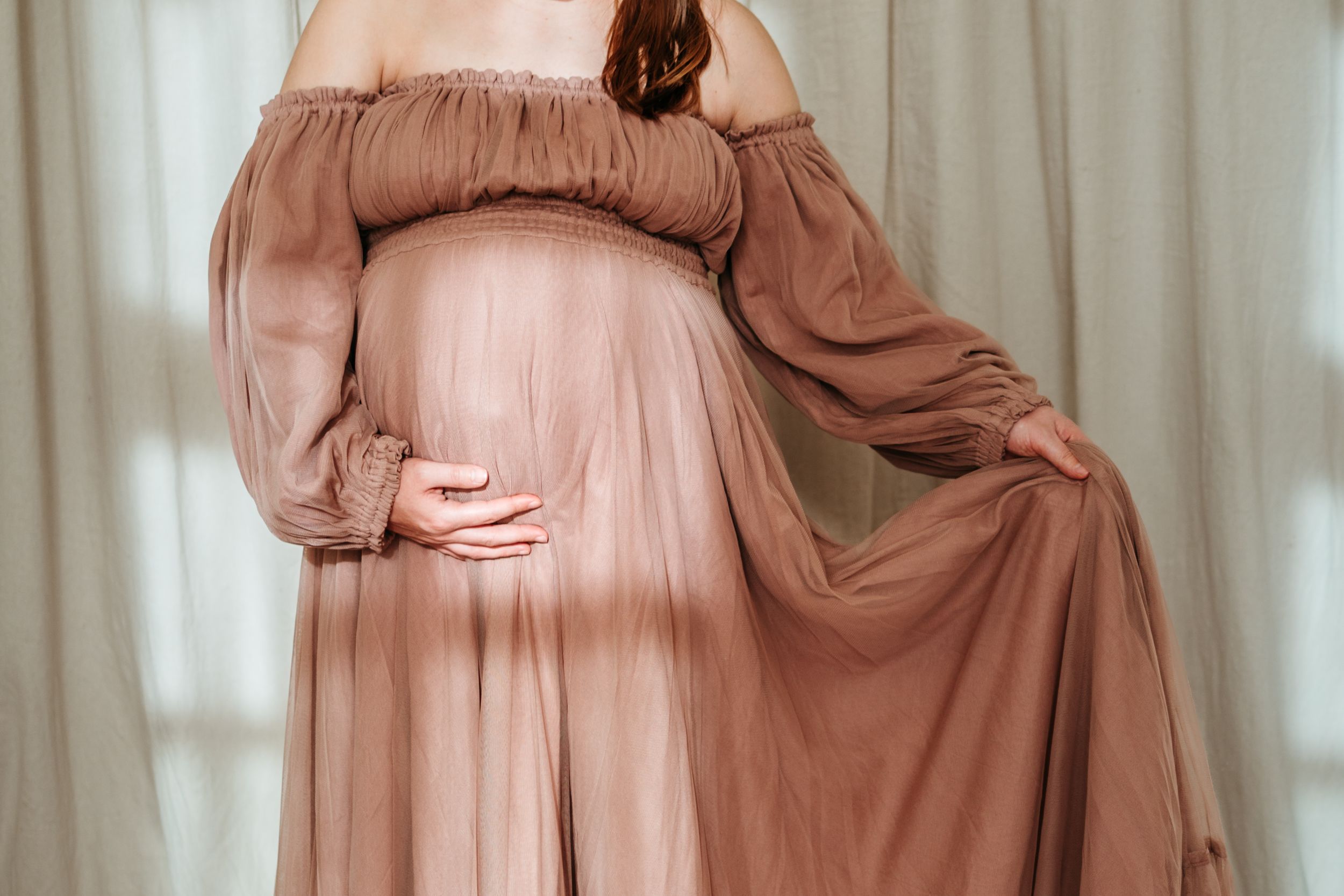 A model poses in a flowing dusty rose gown with delicate draping and elegant movement against a light backdrop.