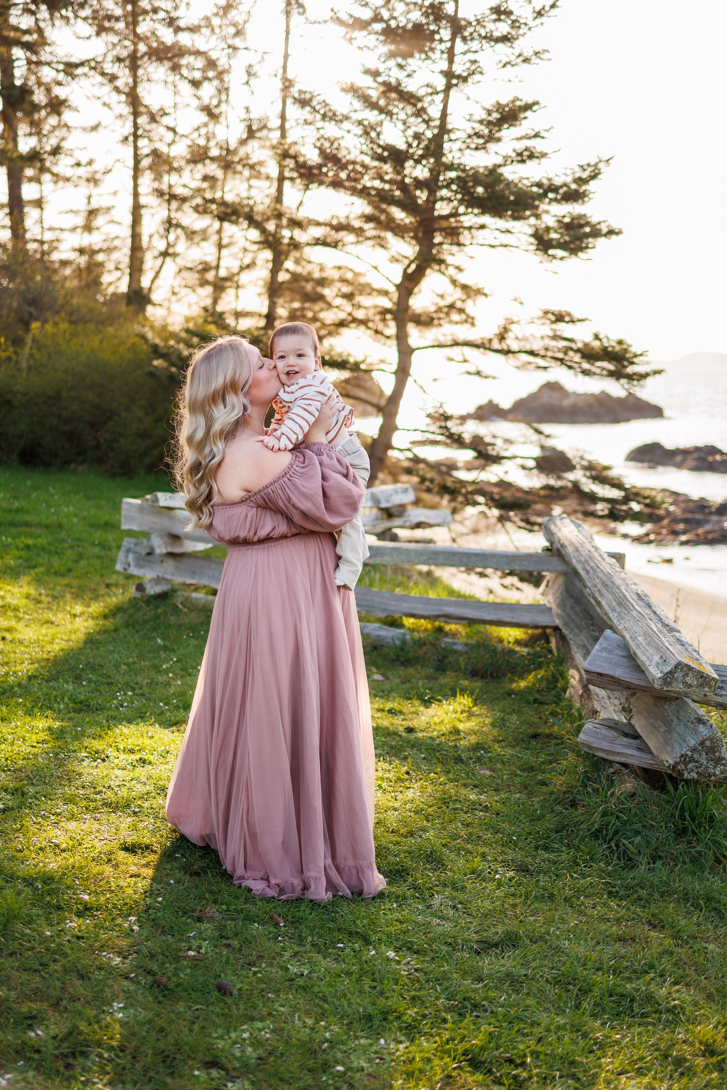 A person in a dusty rose dress holds a baby at sunset near a rocky coastal area with evergreen trees.