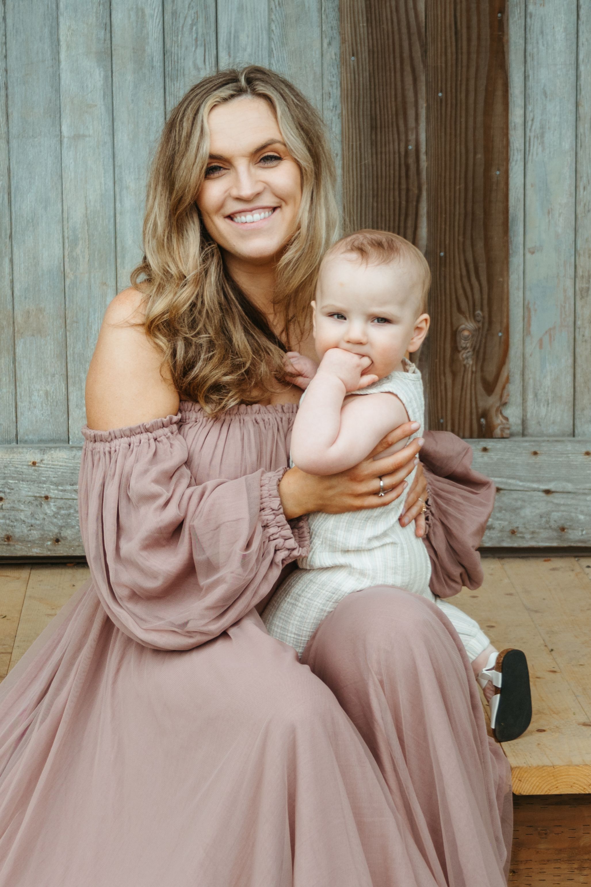 A parent in a mauve off-shoulder dress sits with a baby in white against a rustic wooden backdrop.