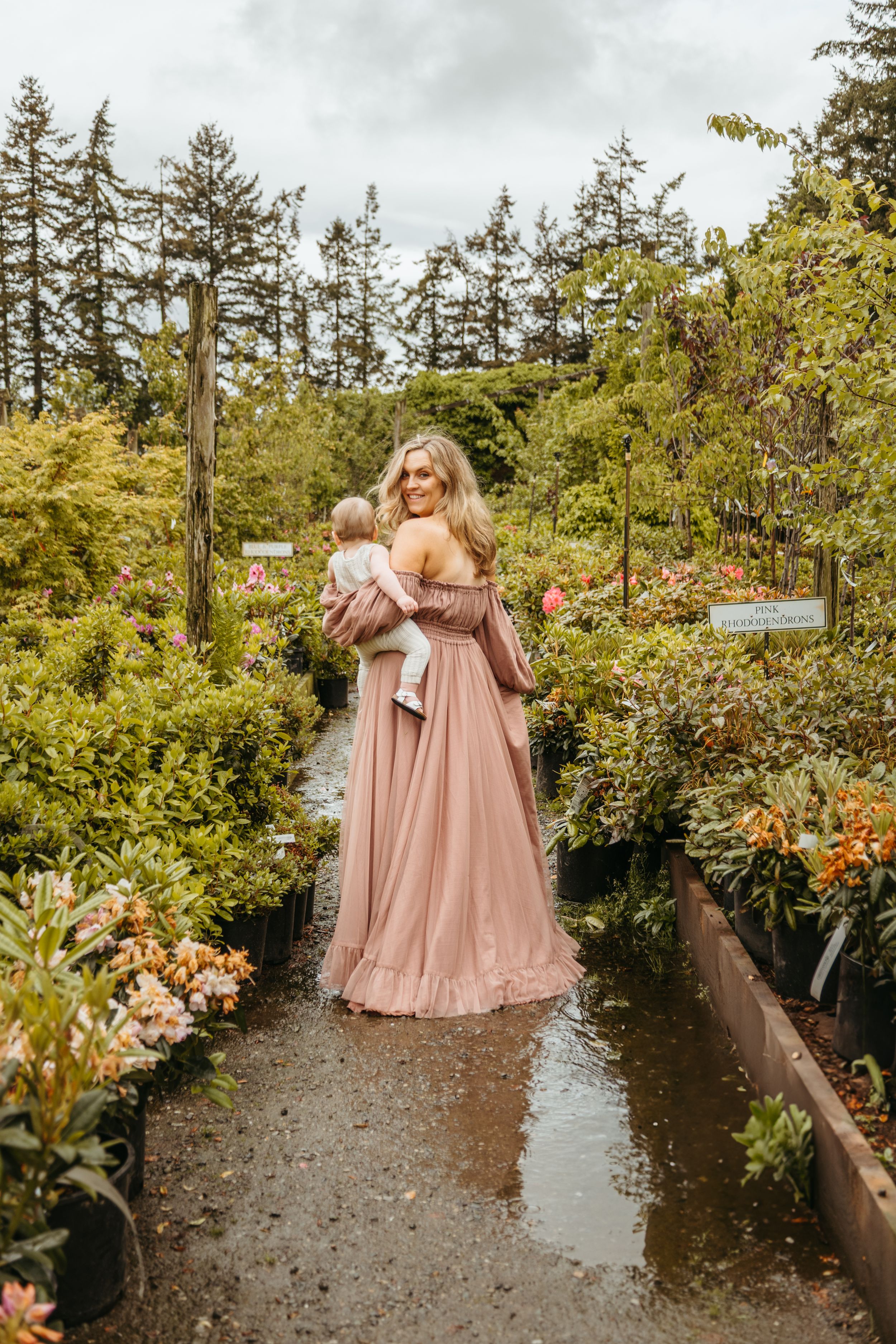 Bridesmaids in flowing blush pink dresses walk along a garden path surrounded by lush greenery and trees.