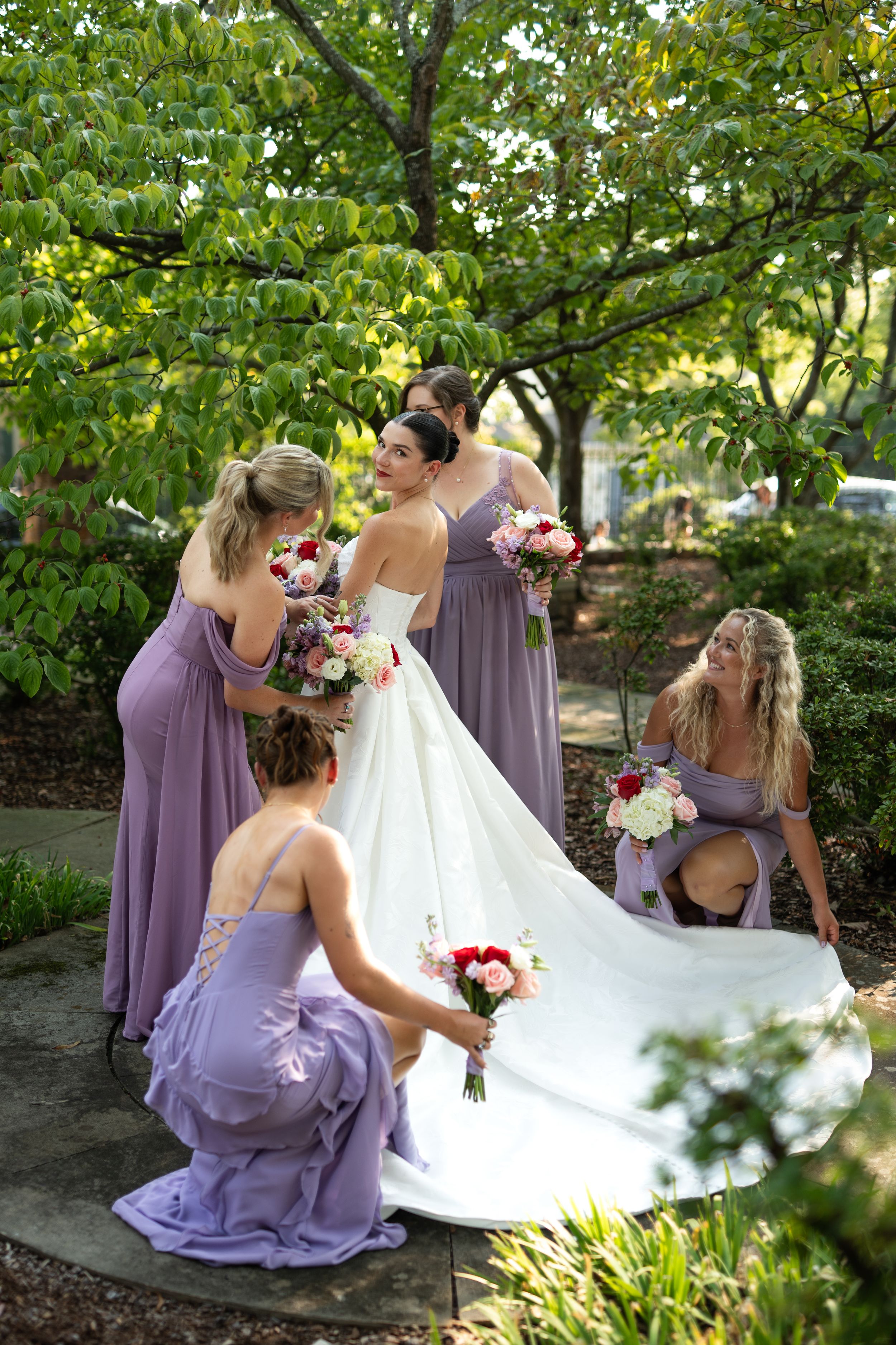 Bridesmaids in lavender dresses helping adjust wedding gown train in garden setting.
