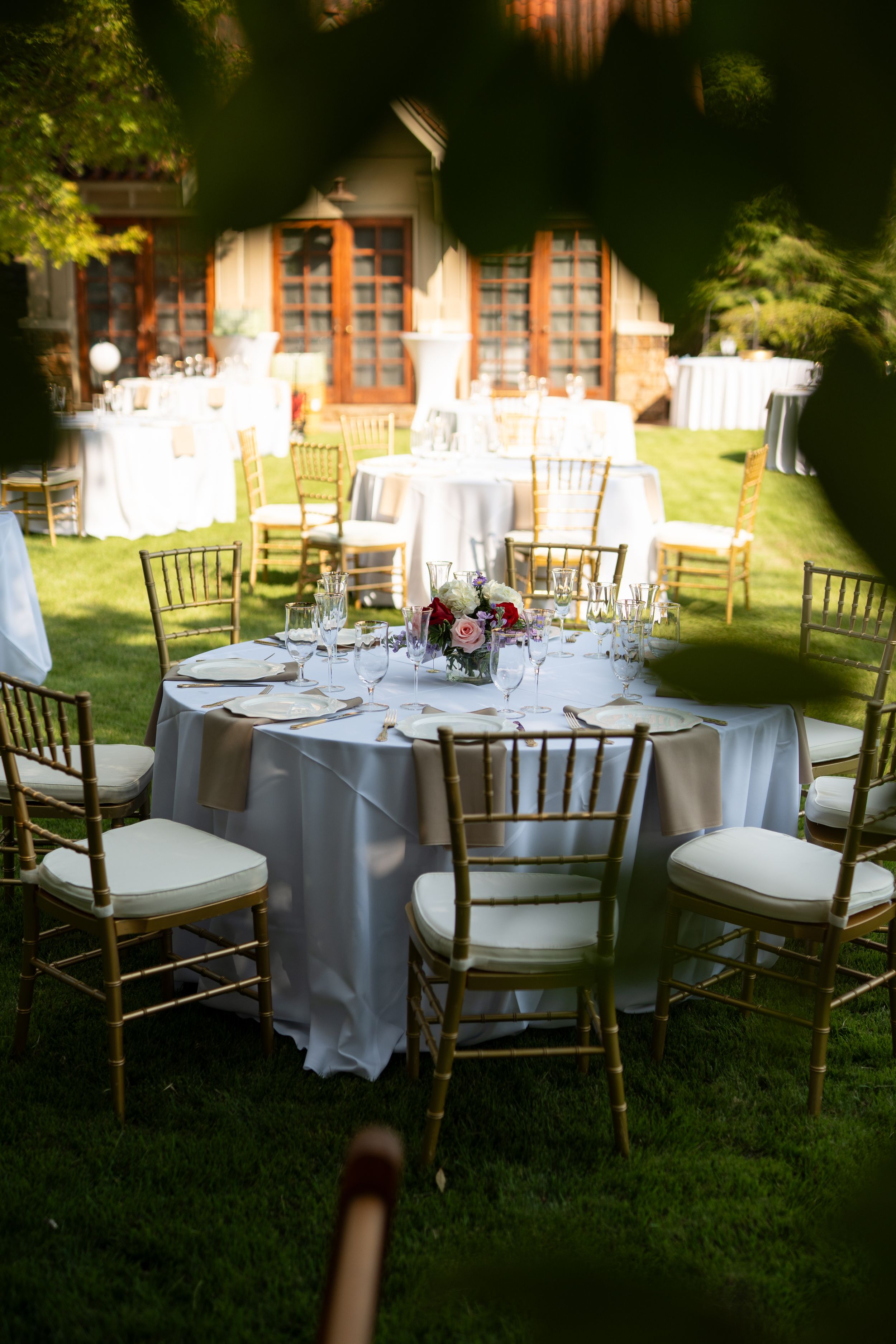 Outdoor wedding reception setup with white tablecloths and wooden Chiavari chairs in garden setting.