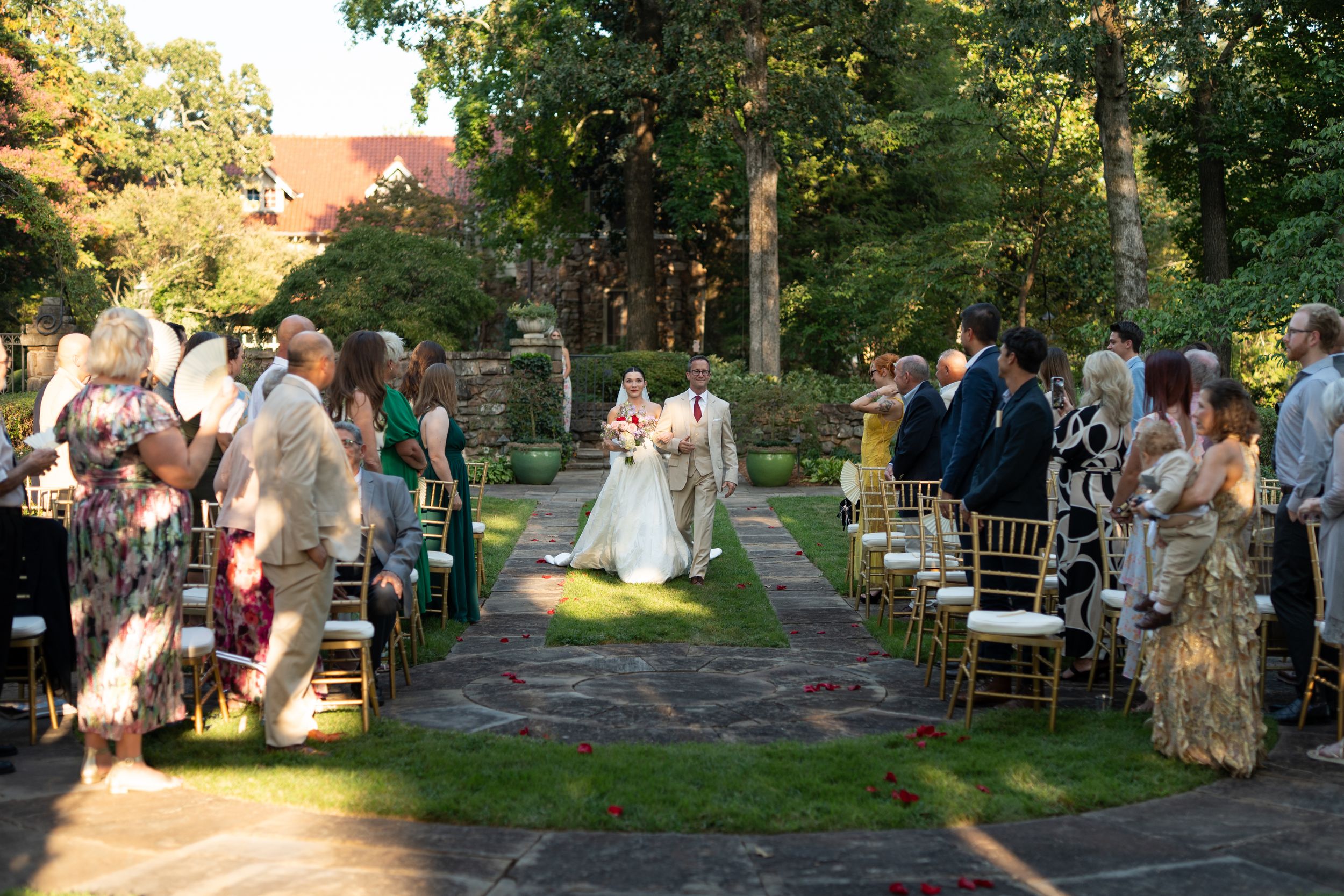 Guests seated outdoors at a garden wedding ceremony with trees and sunlight filtering through.