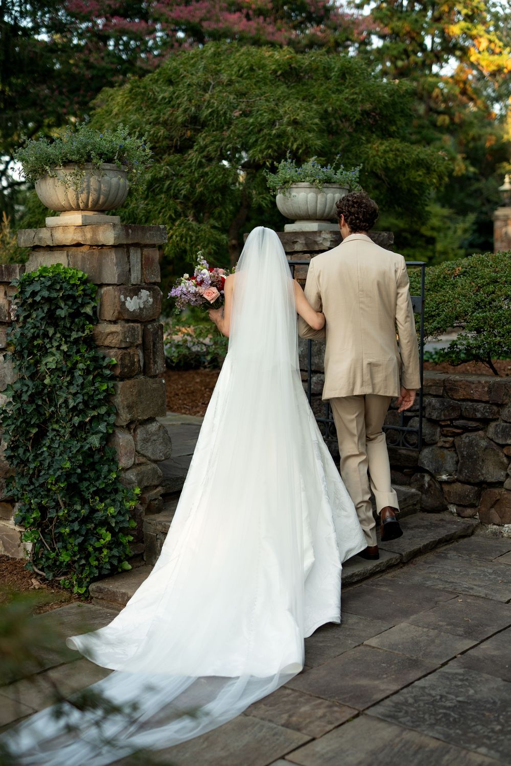 Wedding couple walks along a stone pathway surrounded by lush garden greenery.