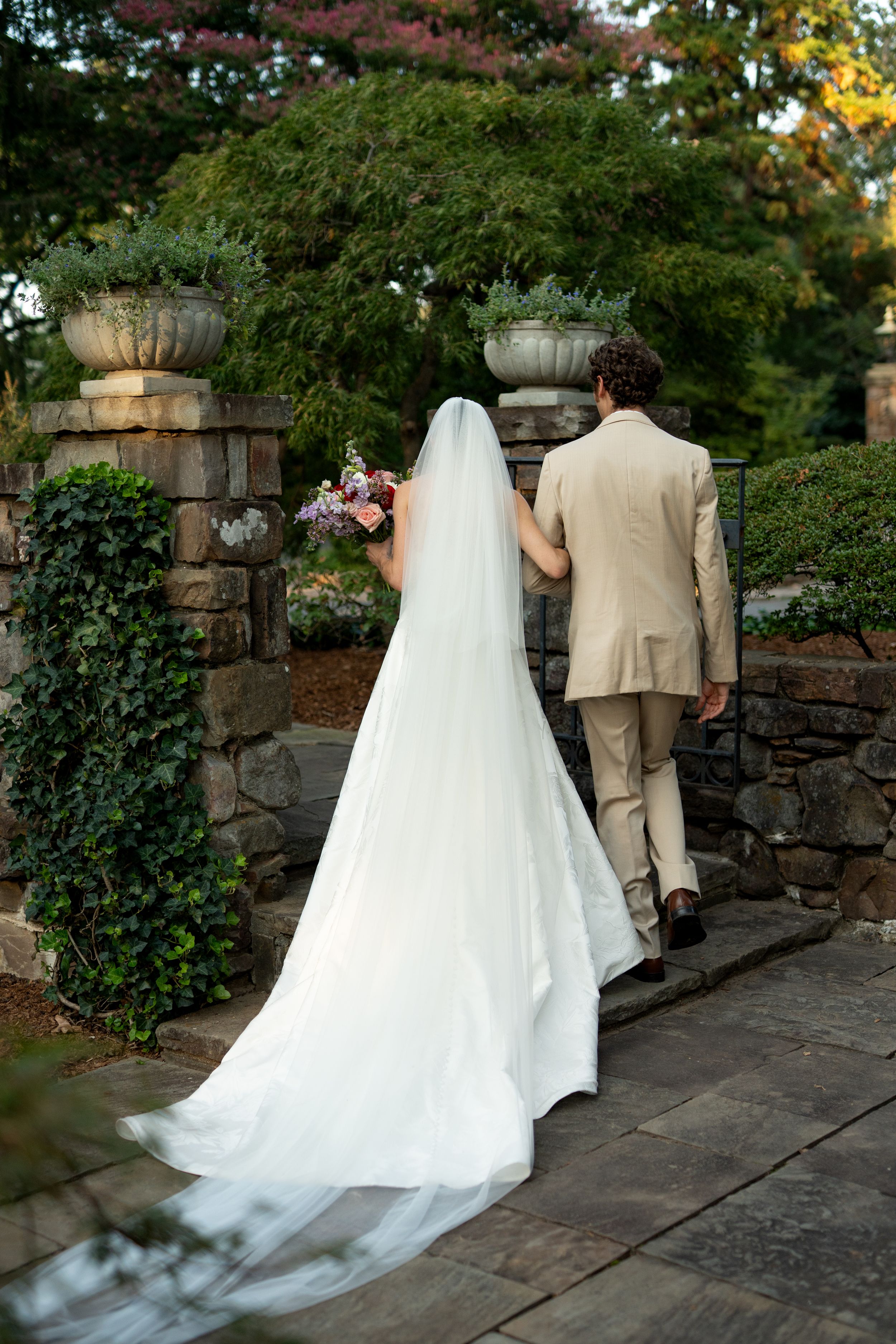 Wedding couple walks along a stone pathway surrounded by lush garden greenery.