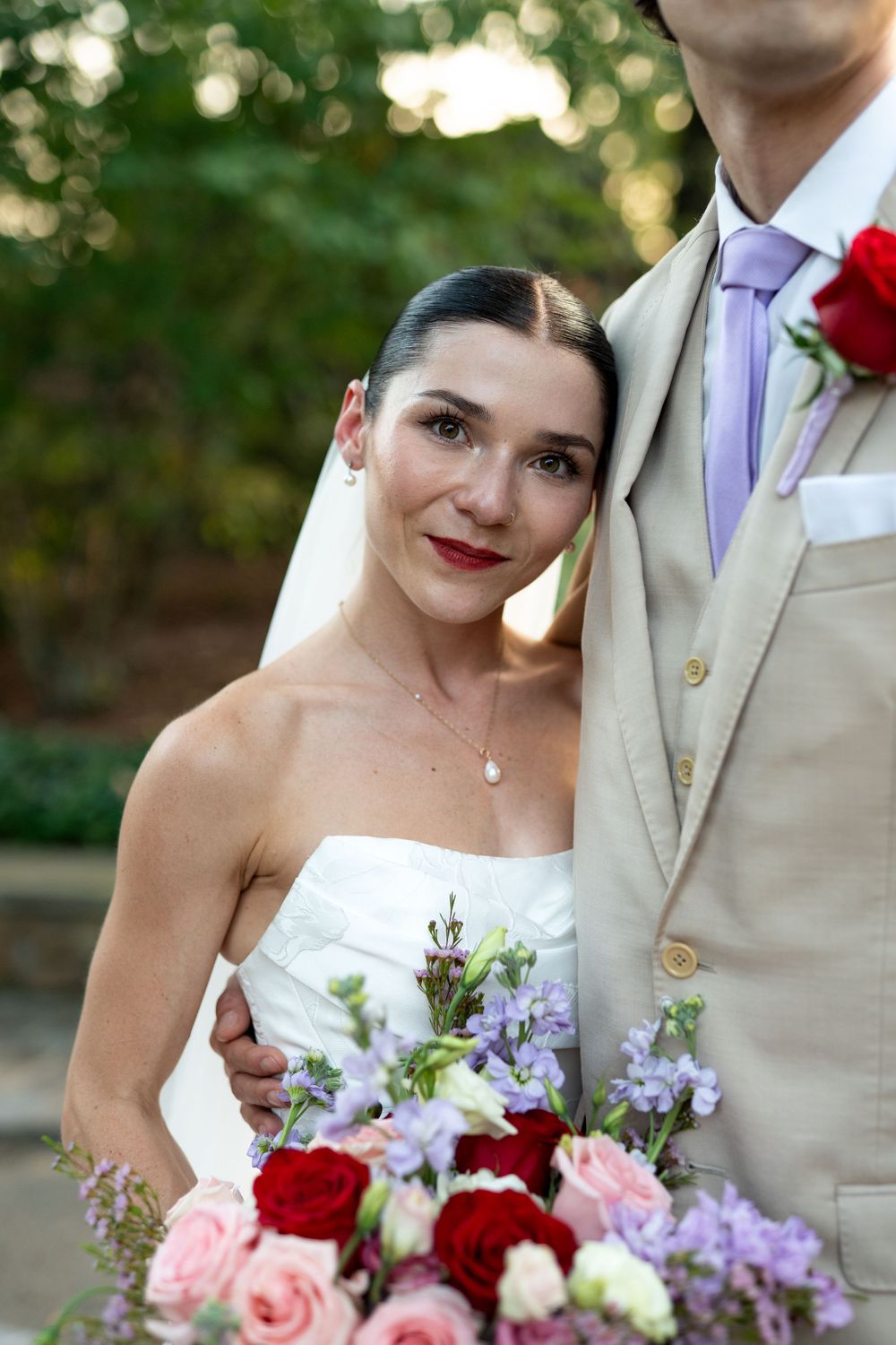 Wedding couple poses with vibrant bouquet featuring red roses and purple flowers.