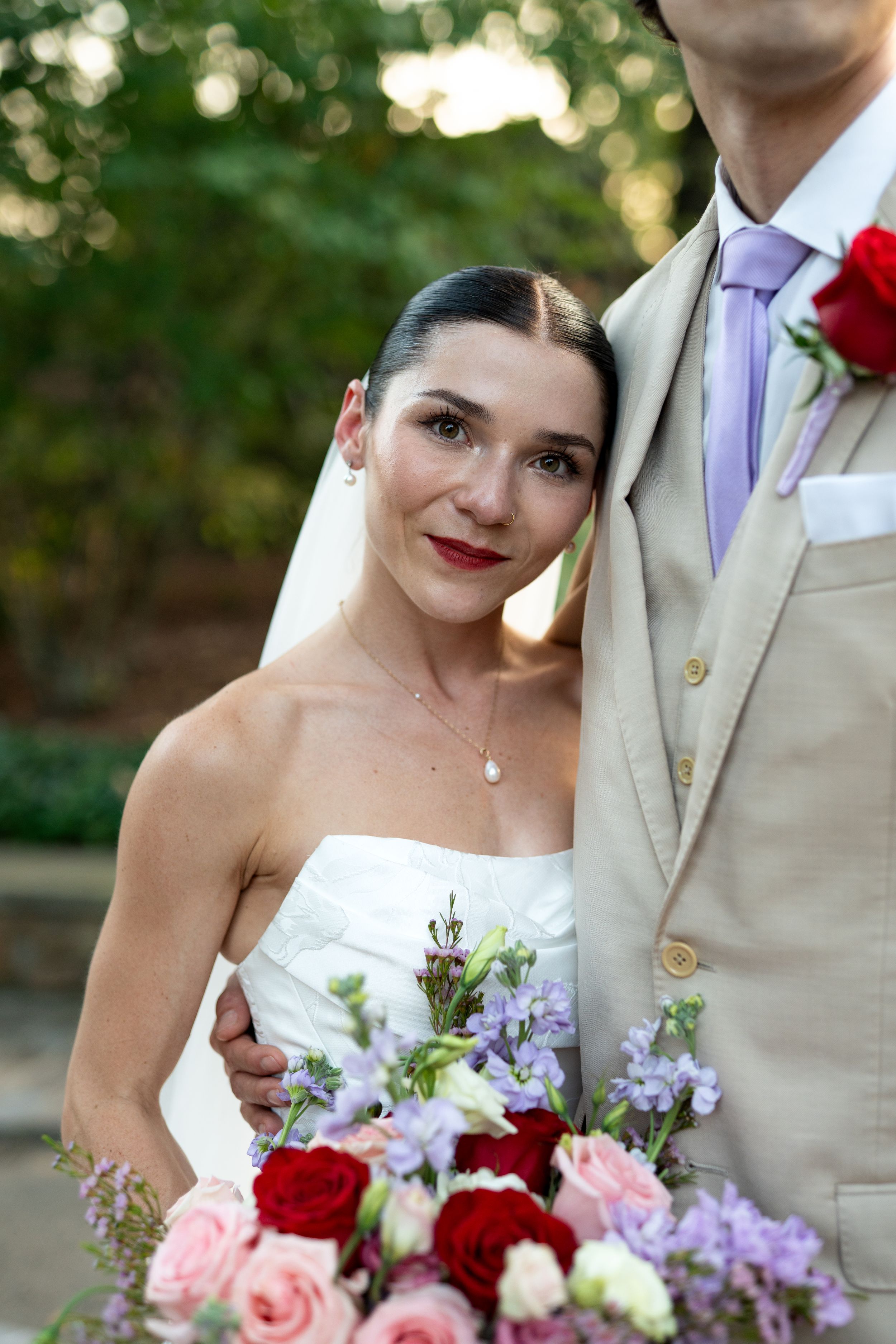 Wedding couple poses with vibrant bouquet featuring red roses and purple flowers.