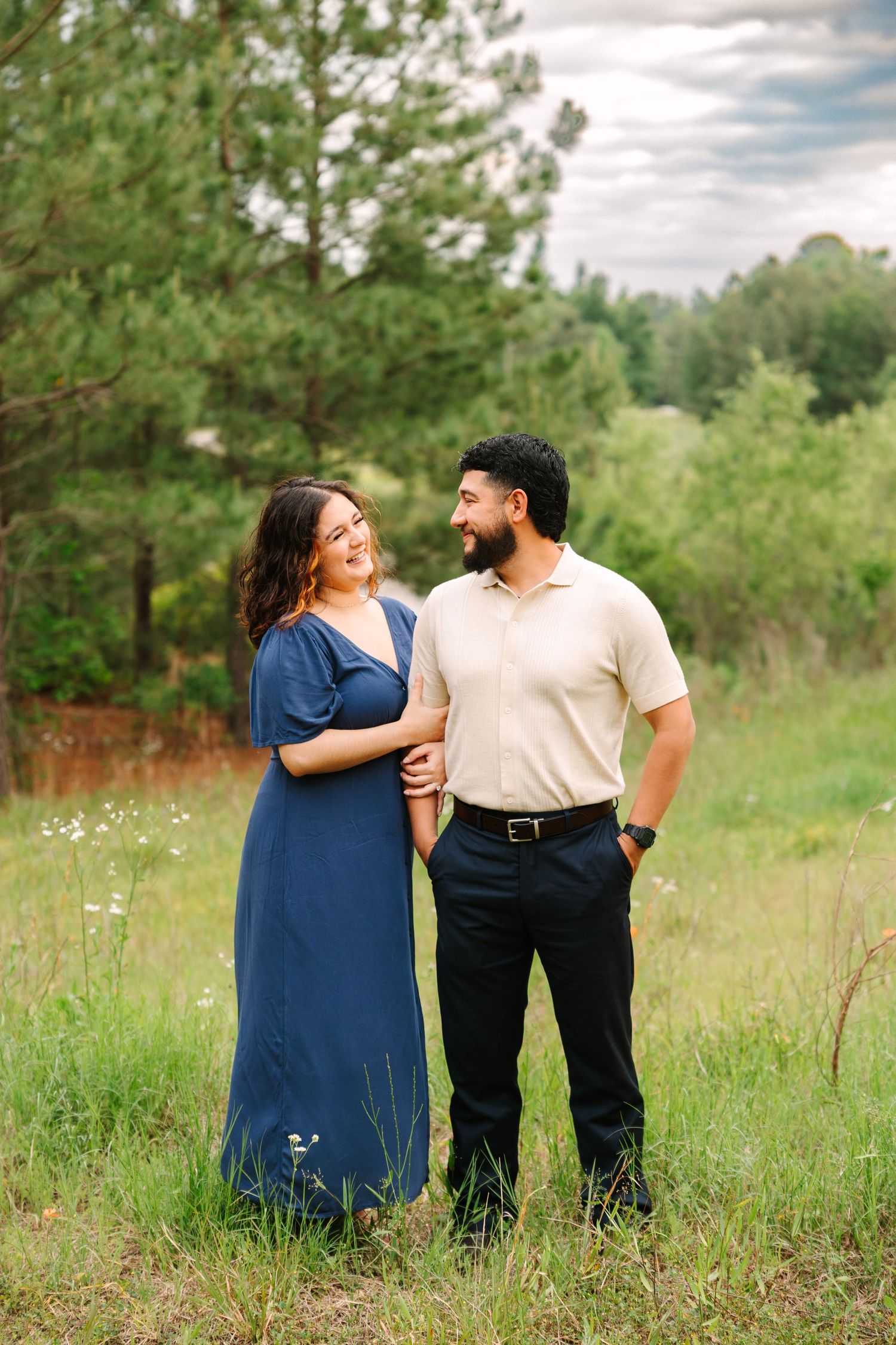 A couple in casual attire shares tender moments together in a sunlit meadow surrounded by pine trees.