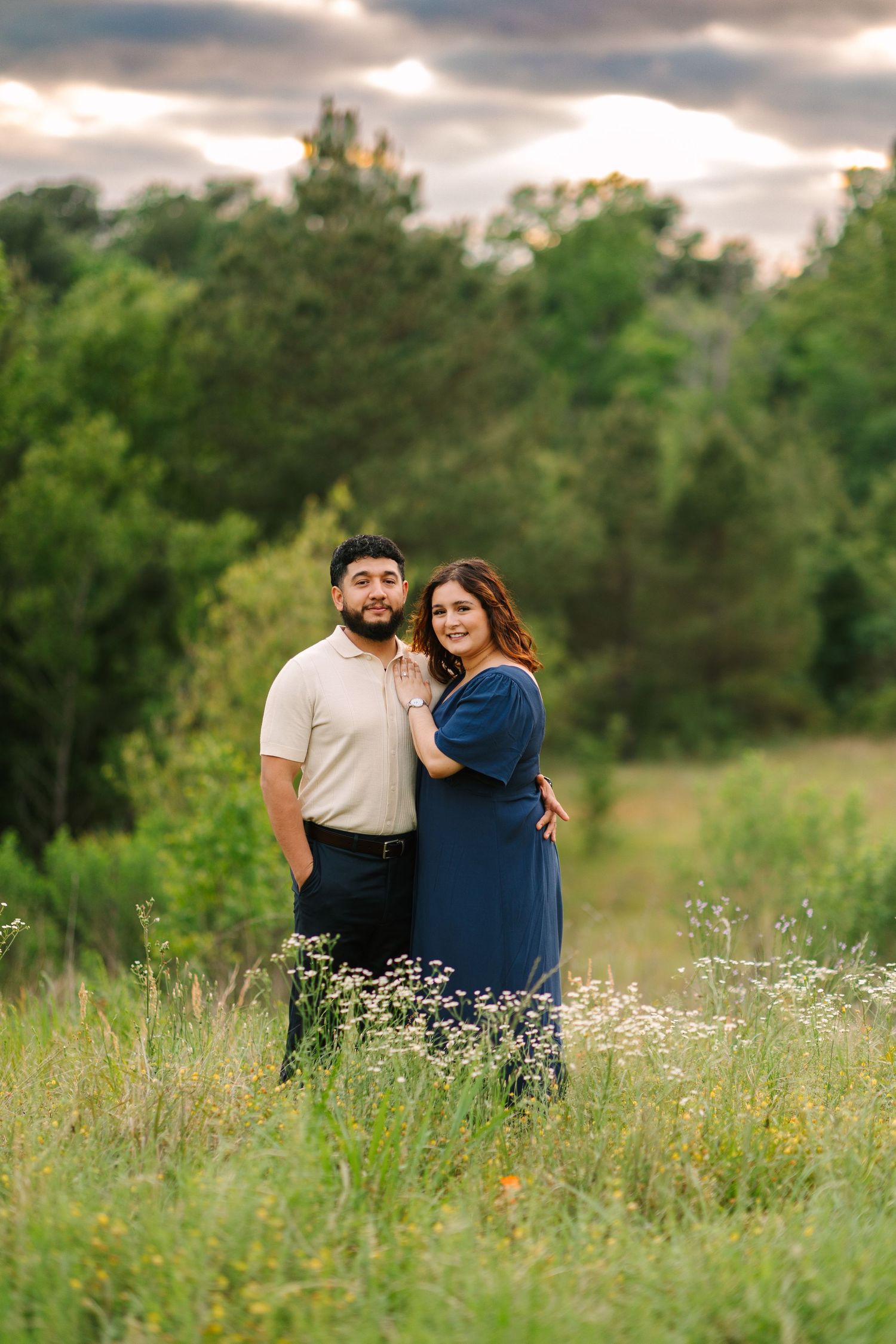 Couple embraces in a sunny meadow with wildflowers and green trees in the background.