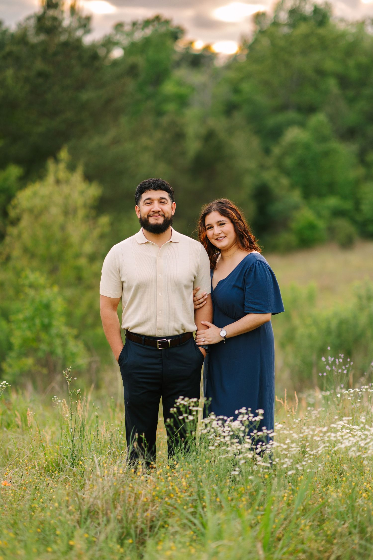 A couple stands together in a wildflower meadow at sunset wearing casual attire.