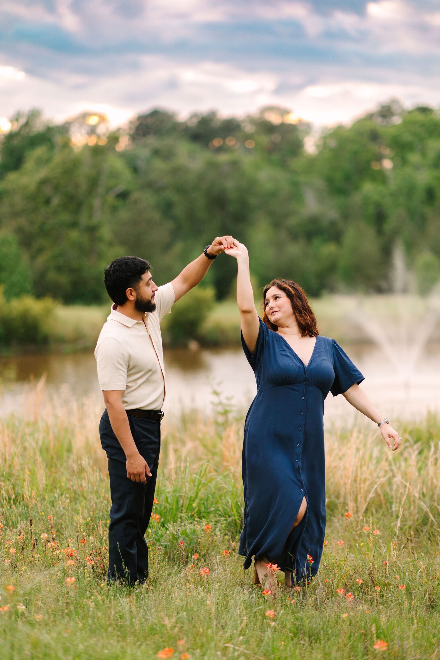 A couple shares a playful moment dancing together in a wildflower meadow near a lake at sunset.
