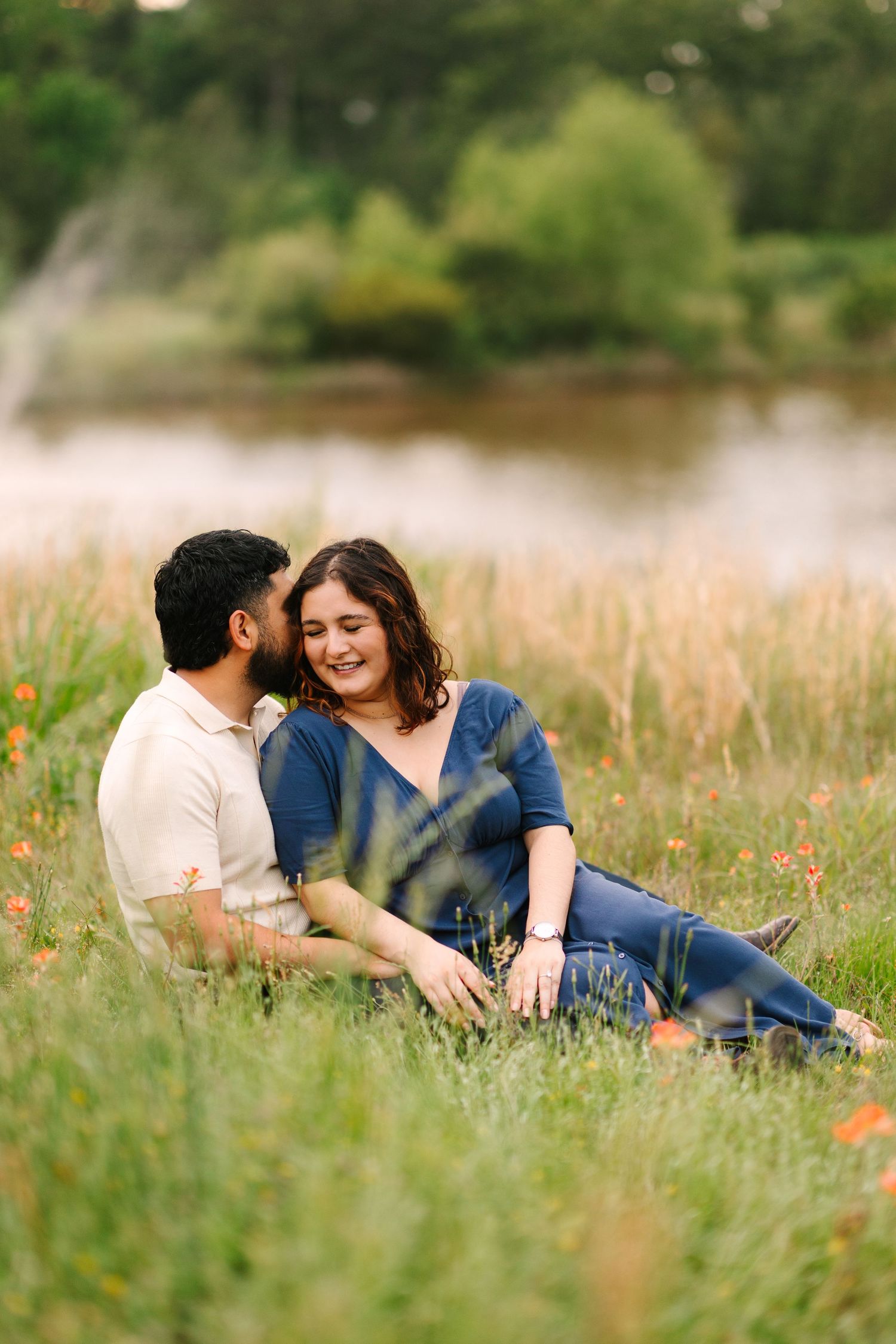 A couple shares an intimate moment while relaxing in a sunlit meadow with tall grass and wildflowers near a pond.