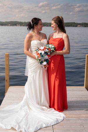 Bride in white gown and attendant in red dress pose together on wooden dock by lake.
