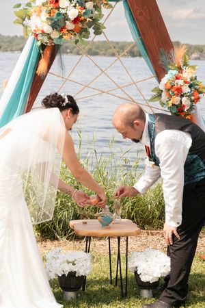 Couple signing marriage documents at lakeside ceremony under decorated wooden arch.