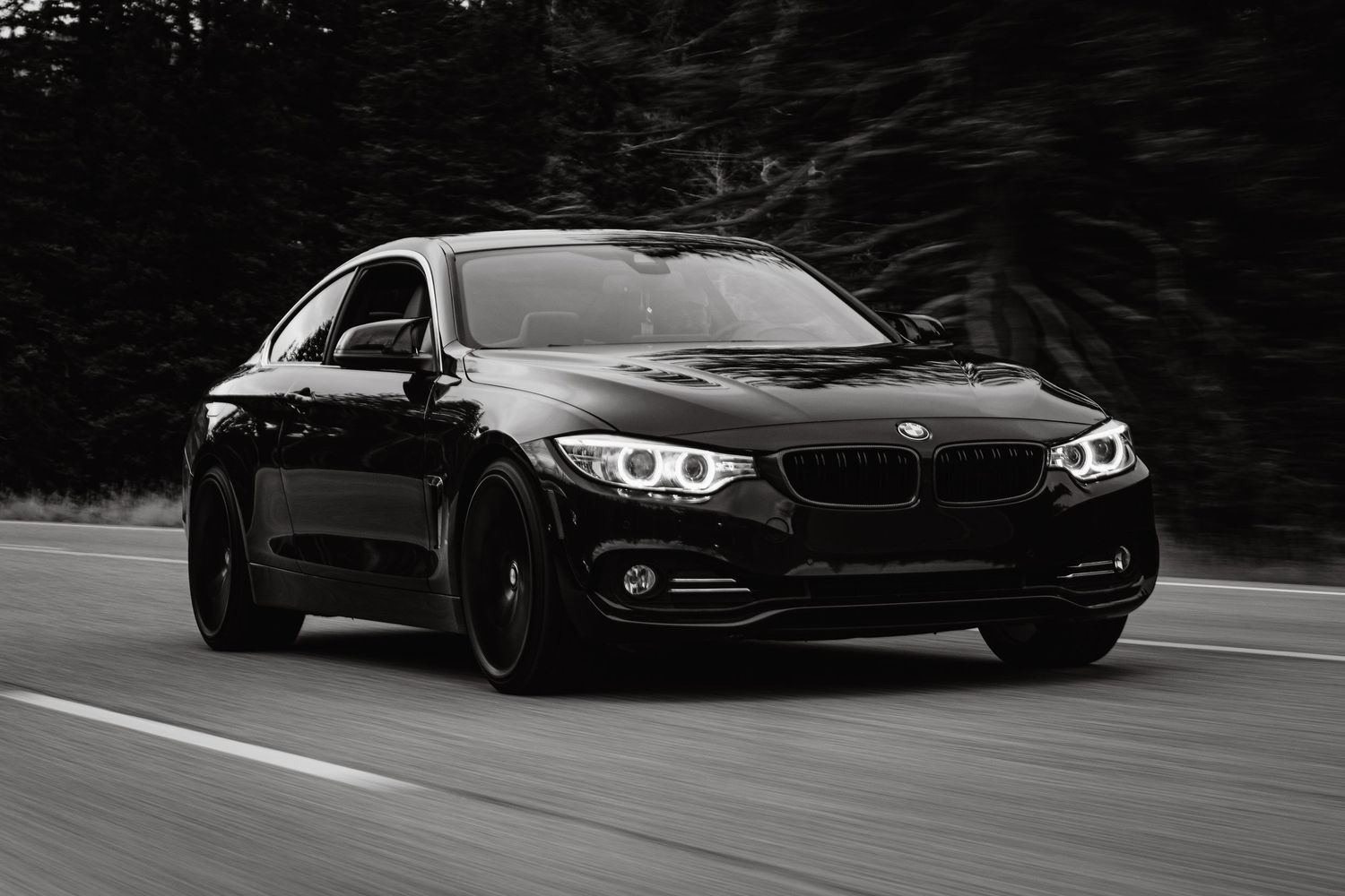 A dramatically lit black and white photo of a black BMW driving down a road.