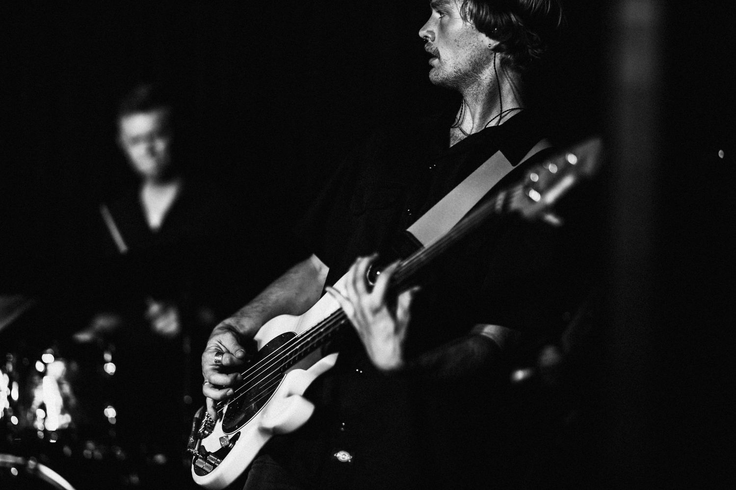A black and white photo of a man playing a bass in a dark room, with a blurry drummer in the background.