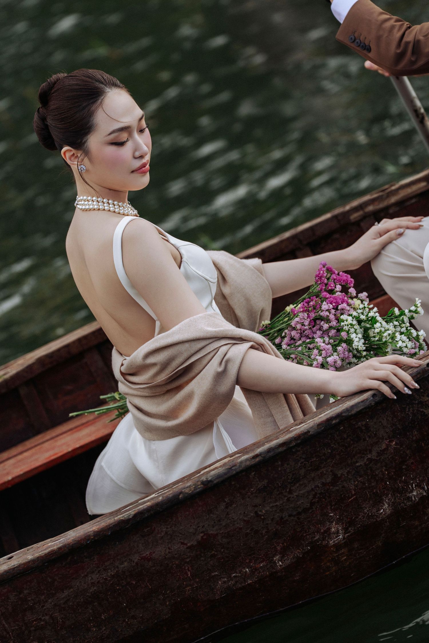 Bride in white dress with beige wrap holding flowers while seated in a wooden boat on a calm lake.
